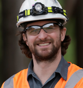 A man wearing a hard hat, reflective vest, and safety glasses smiles