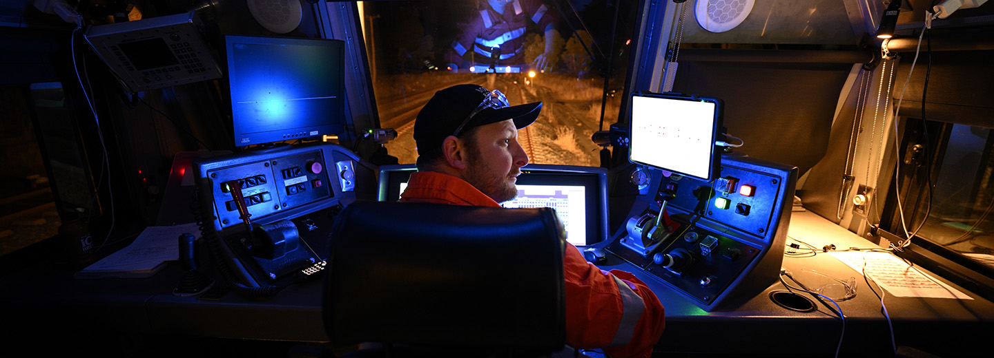 A man in a baseball cap sits at a dashboard watching a series of screens.