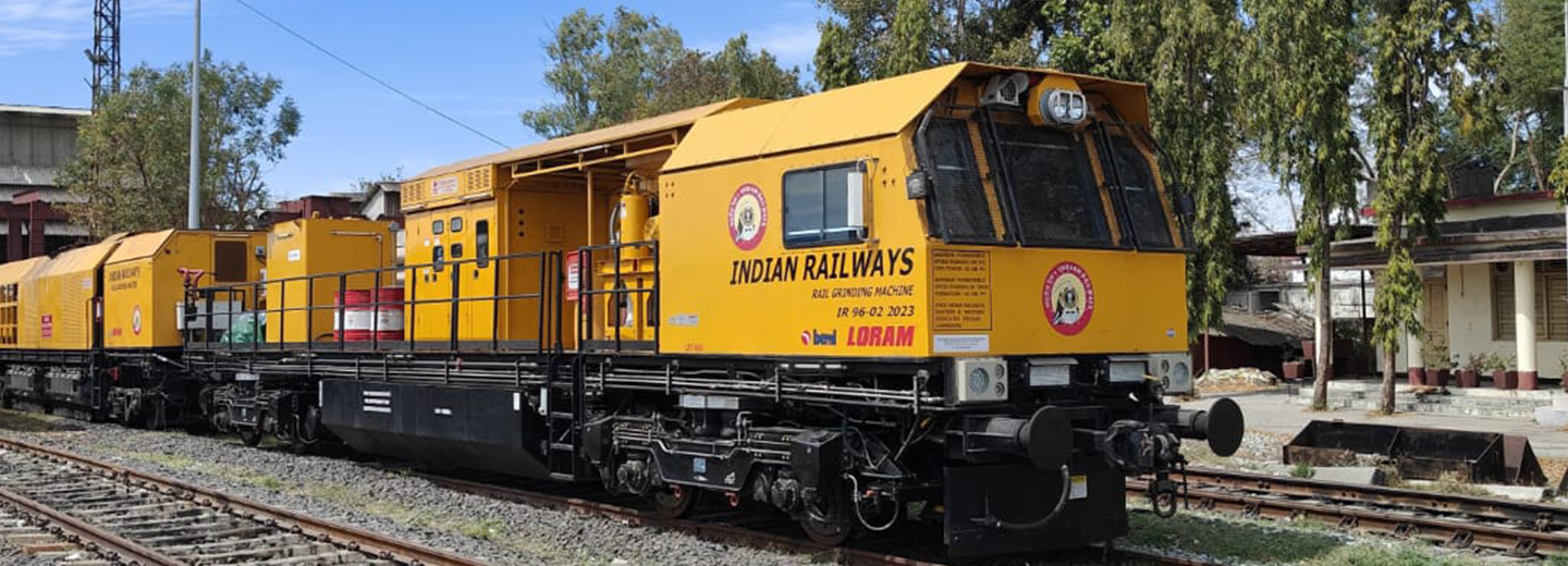 A yellow train on tracks outside in front of trees under a blue sky.